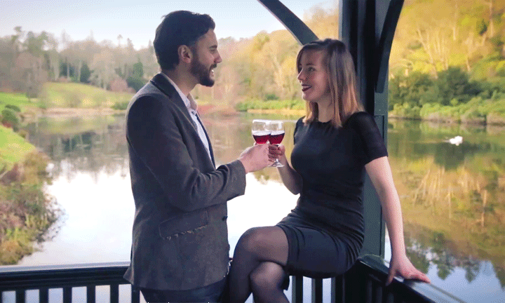 A couple raise their glasses of wine on a balcony overlooking a loch.